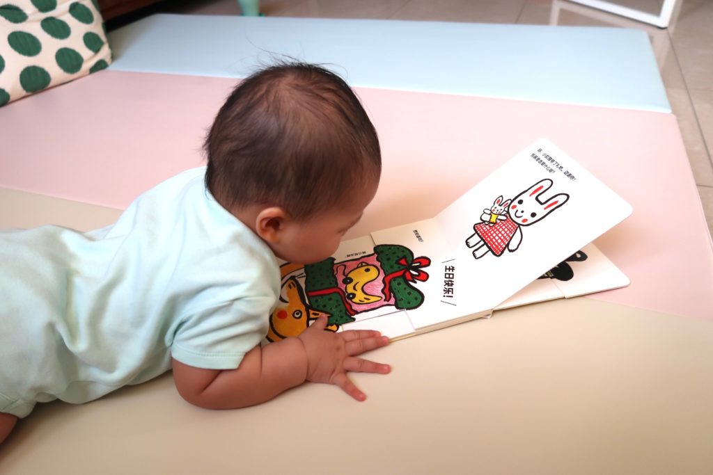 A baby is lying on his belly on a pastel-coloured mat. He pushes up on his arms to examine a board book, which shows a picture of a rabbit in a pink dress.